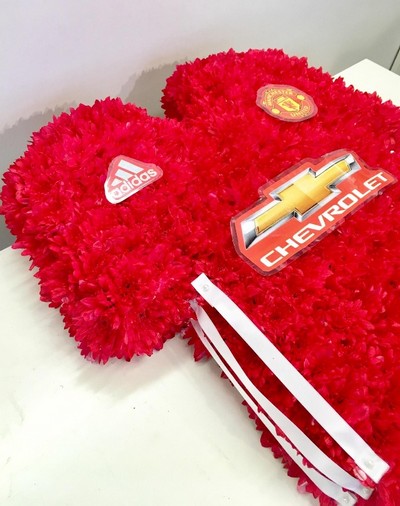 Manchester United shirt funeral tribute with fresh flower base, pipework detailing and printed waterproof badges, 21 x 21 inches, available in classic and modern shirt versions.