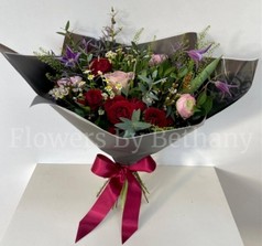 Hand-tied bouquet featuring ranunculus, roses, matricaria, clematis, and seasonal florals, presented in signature black wrap with a satin bow.